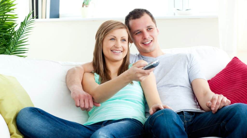 A man and woman sit on a white sofa; she points a remote control forward.