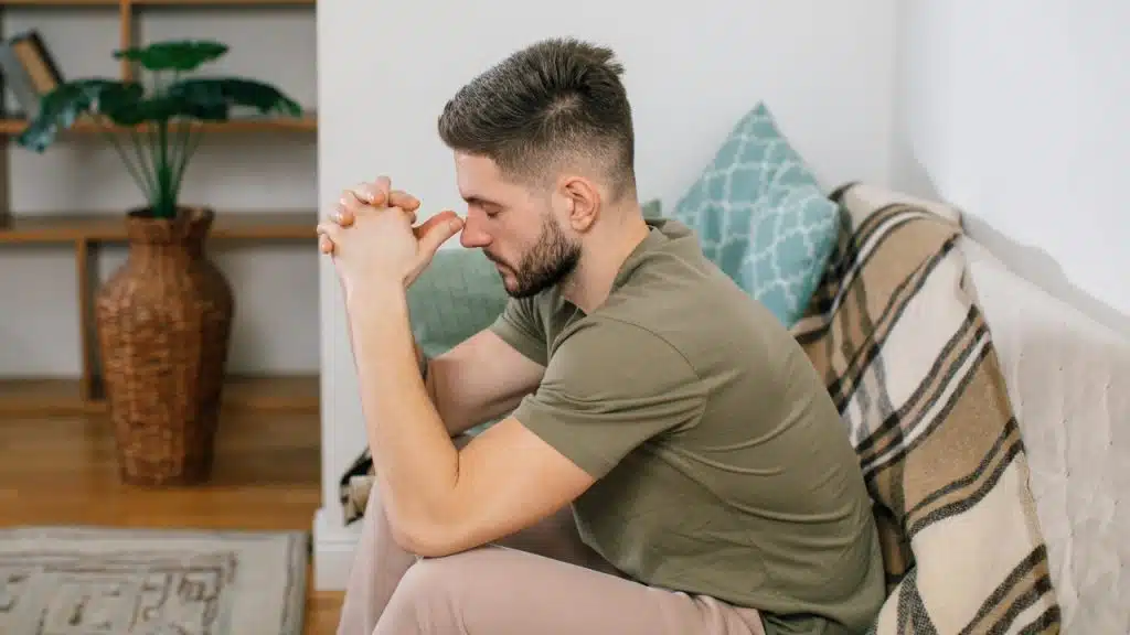 A man with a beard sits on a sofa with his hands clasped near his forehead.