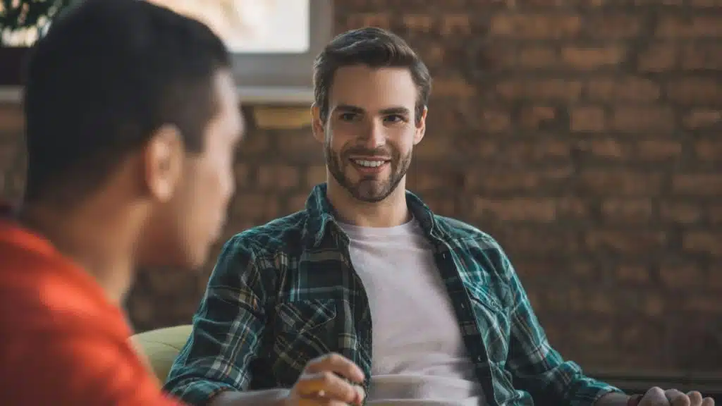 A man in a plaid shirt smiles while looking at someone off-camera during a conversation.