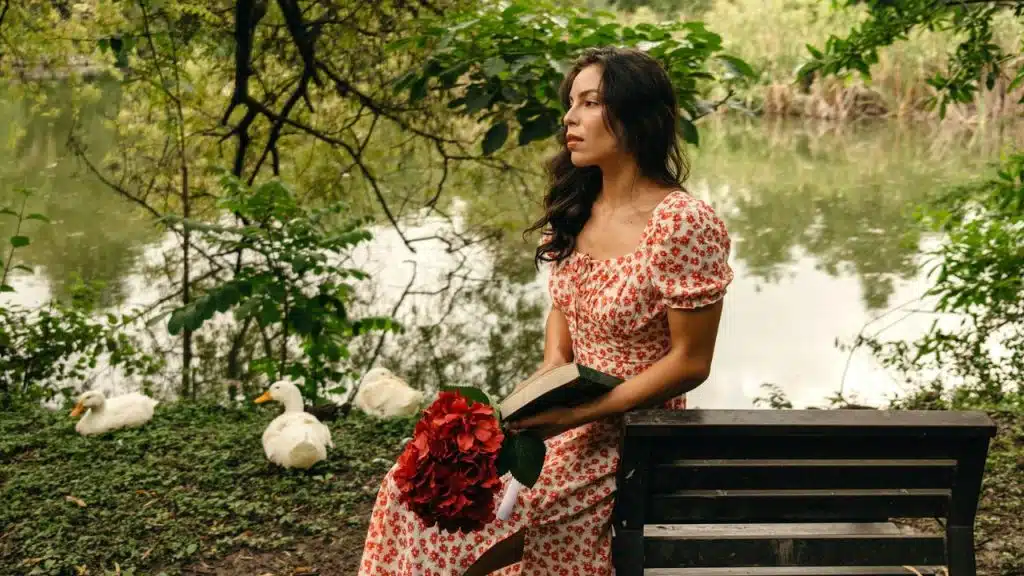 A woman in a floral dress sits on a bench holding a book and flowers.