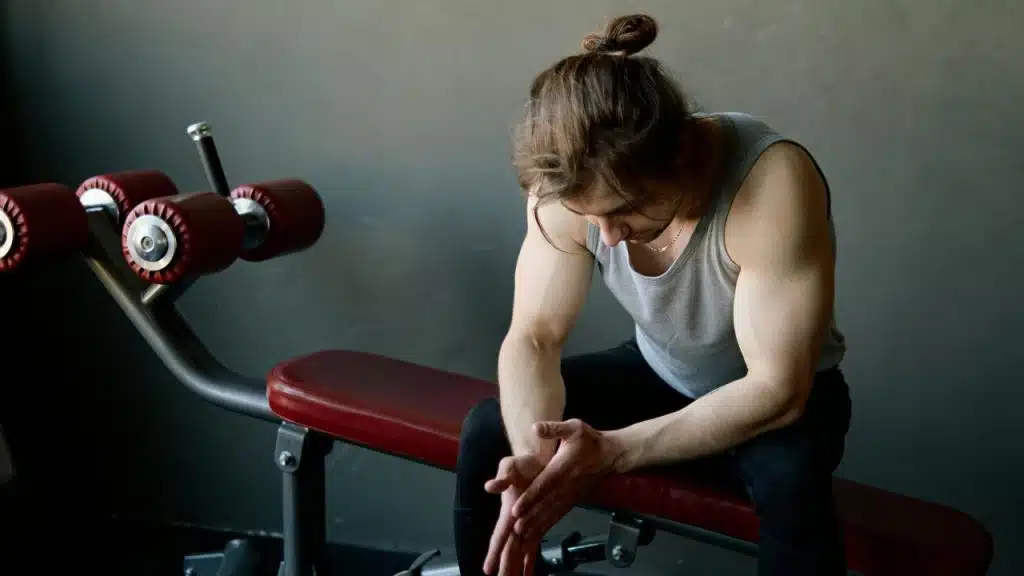 A man with long hair tied back sits on a weight bench with his head down.