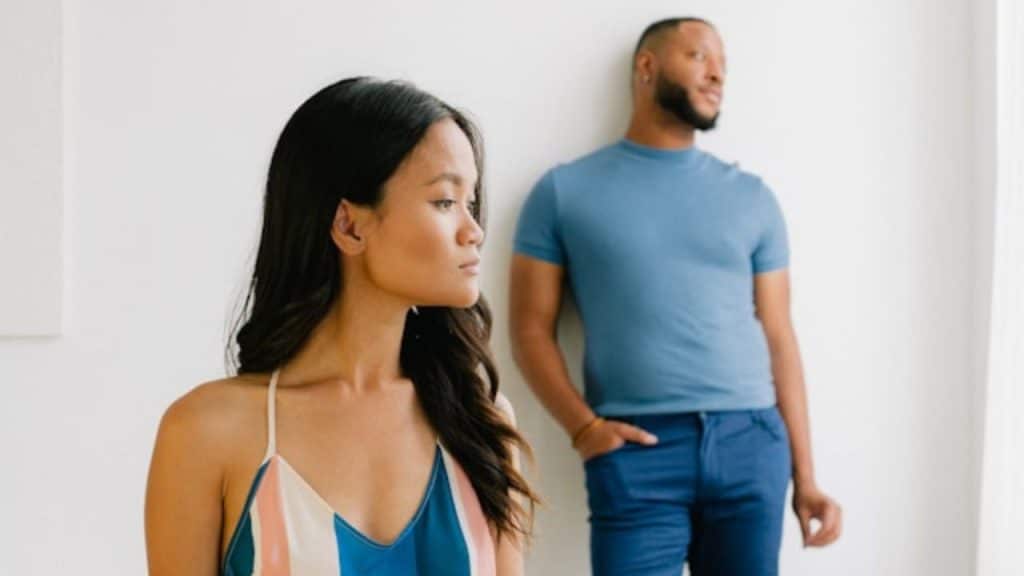 A woman looks away while a man in a blue shirt leans against a wall.