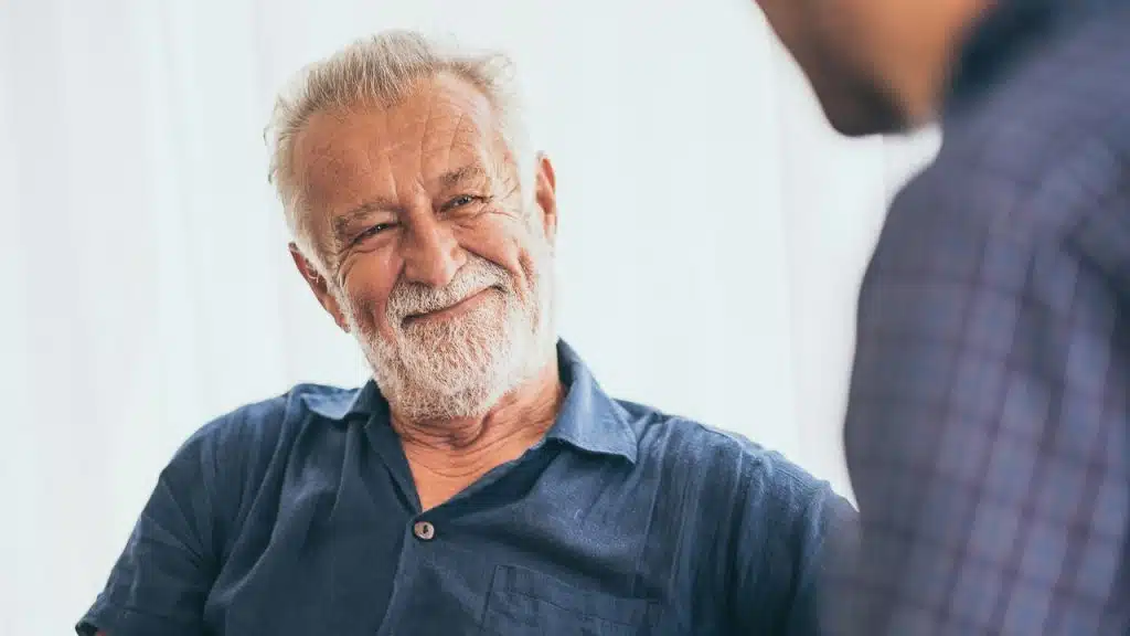An older man with a white beard and grey hair smiles while looking toward someone.