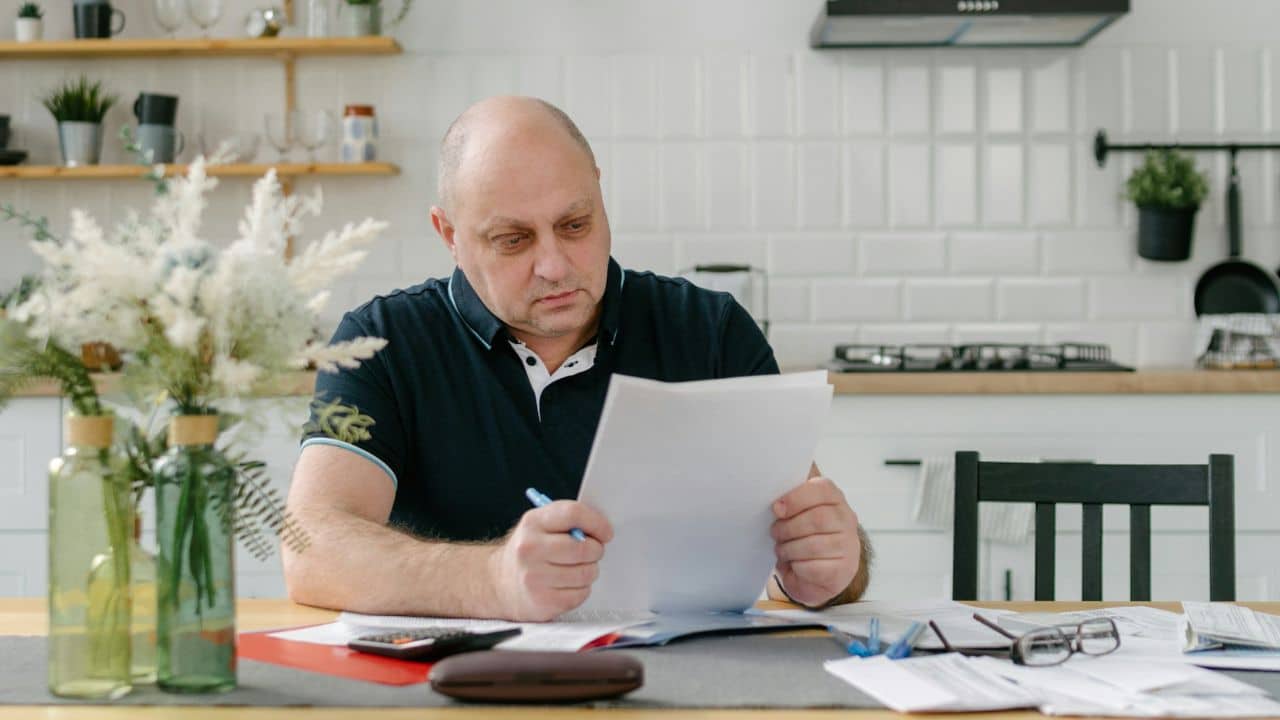 A man sits at a kitchen table while reviewing and holding several white paper documents.