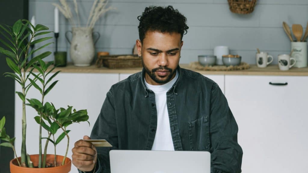 A man with curly hair holds a gold card while looking at a laptop screen.
