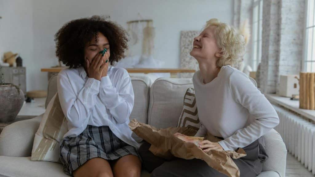 A pair of women laughing on a couch, one holding a wrapped gift.