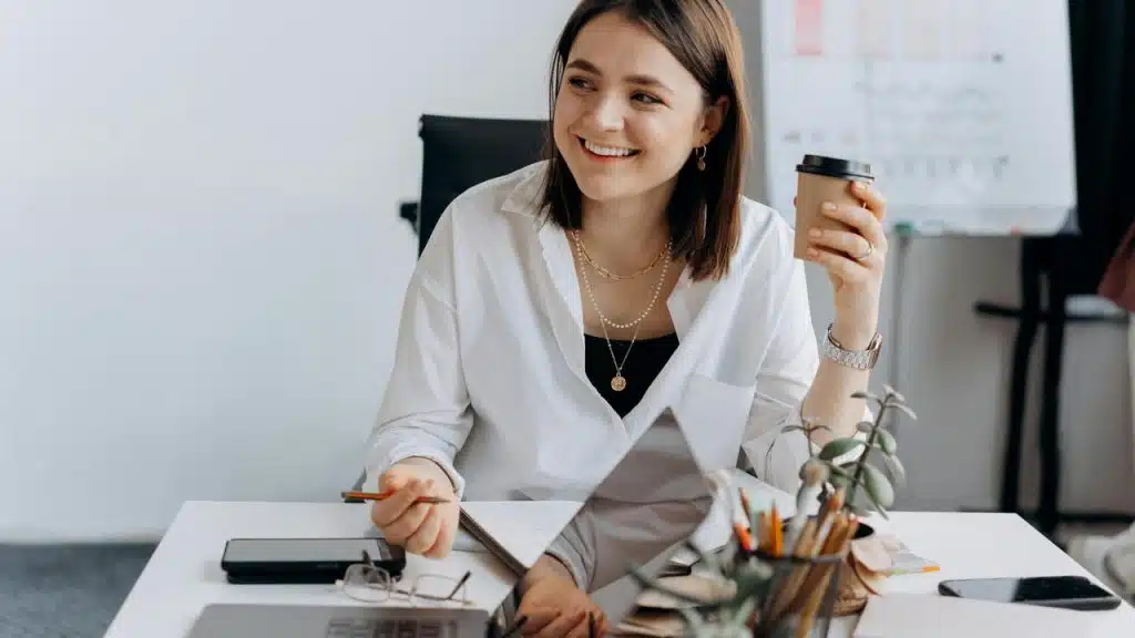 A smiling woman holding a coffee cup while working at a desk.