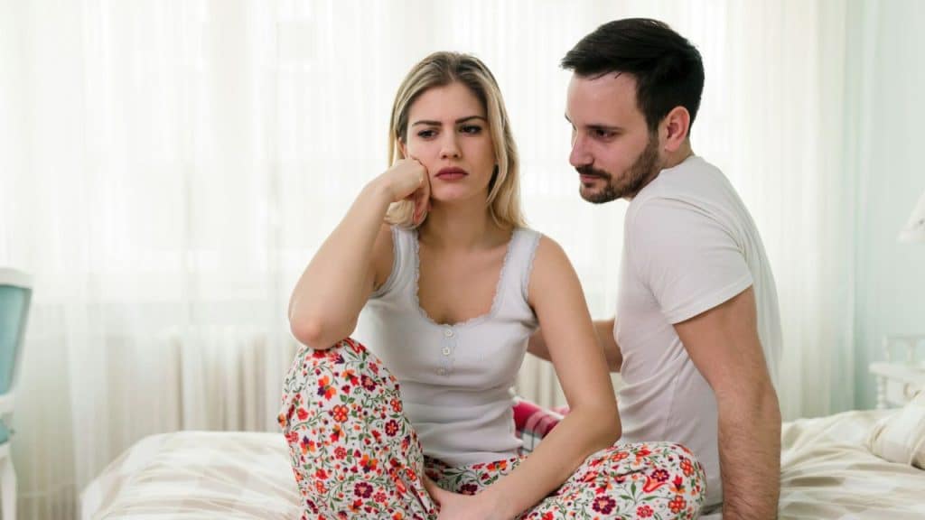 A woman sits on a bed with a hand on her chin while a man watches.