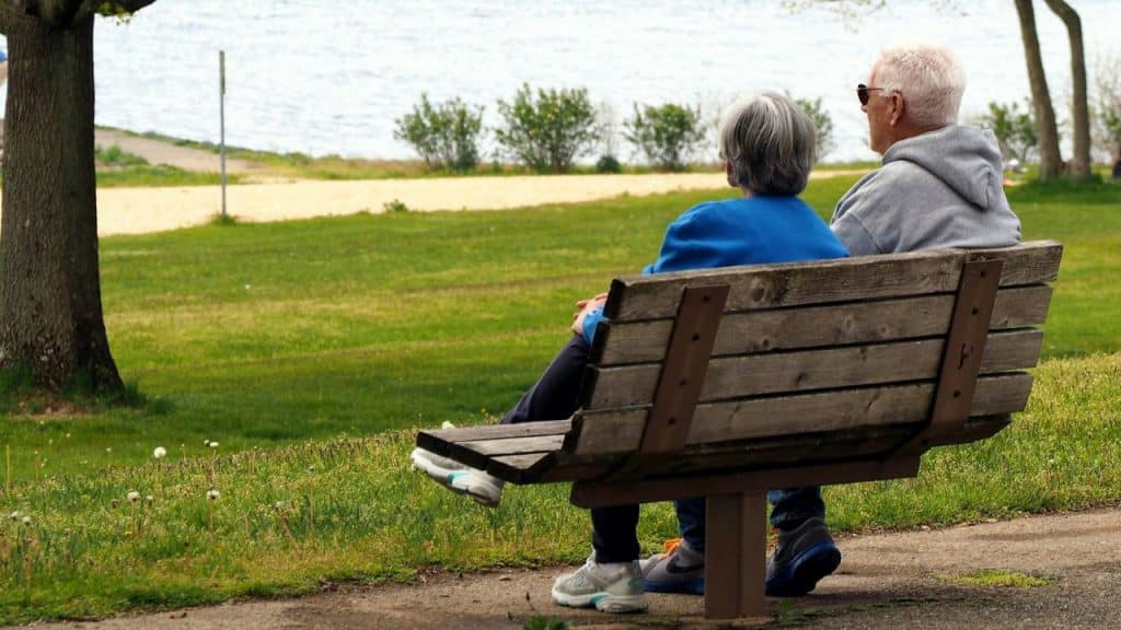 An older couple sits on a wooden park bench looking out toward a body of water.