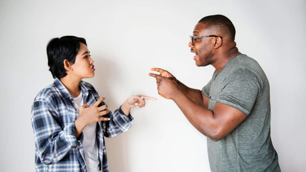 A man and woman stand against a white wall gesturing with their hands while speaking.