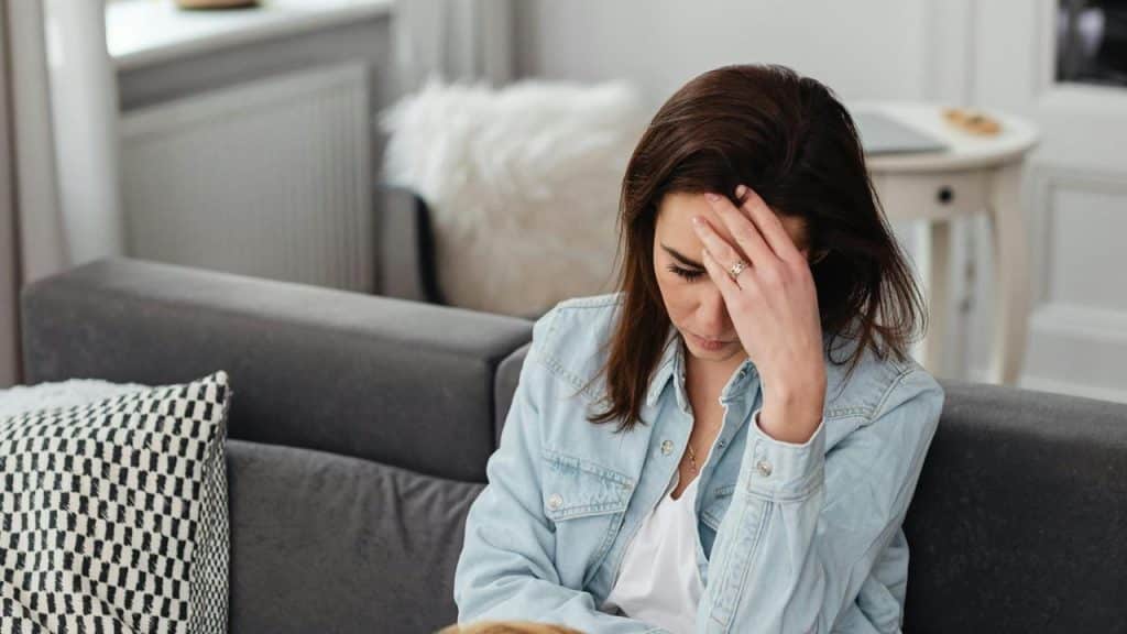 A worried woman sitting on a couch with her hand on her forehead.