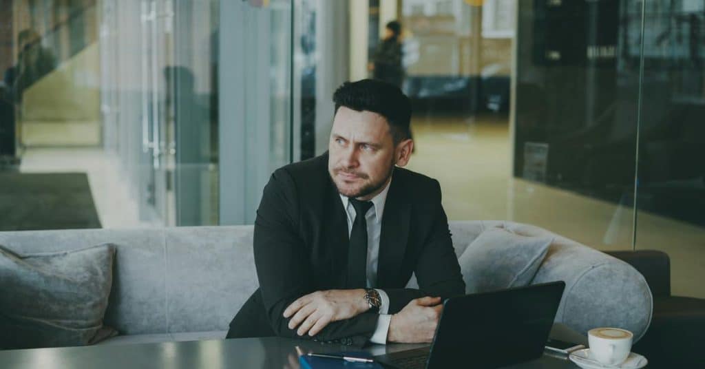 A businessman in a suit sitting with a laptop and looking thoughtfully to the side.