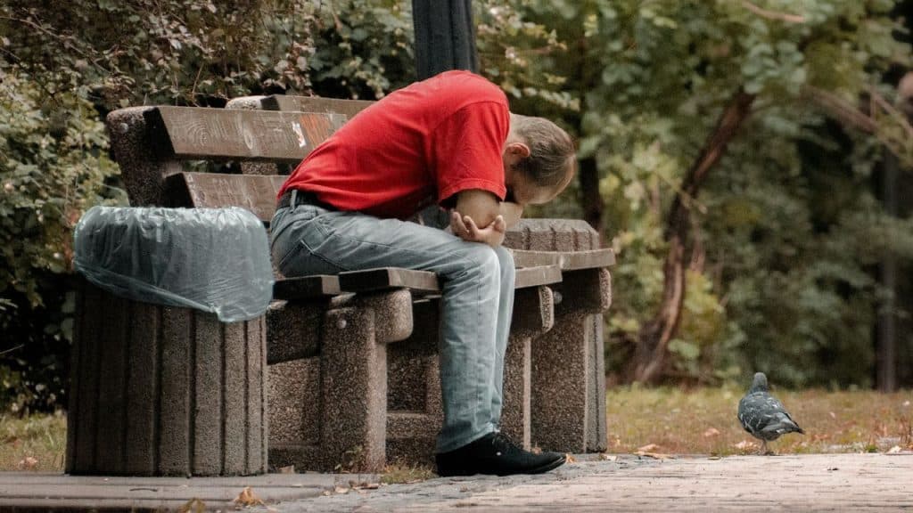 A man in a red shirt sits on a park bench with his head down low.