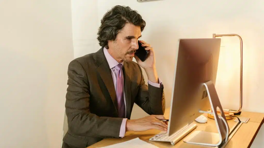 A man in a suit talks on a smartphone while sitting at a computer desk.