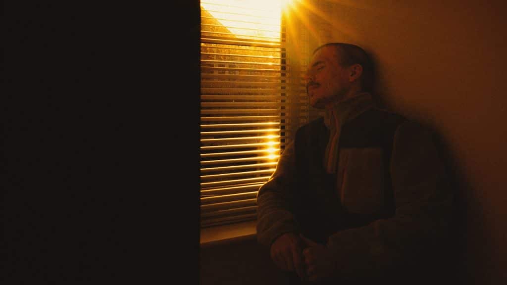 A man sits against a wall in a dark room with sunlight streaming through window blinds.