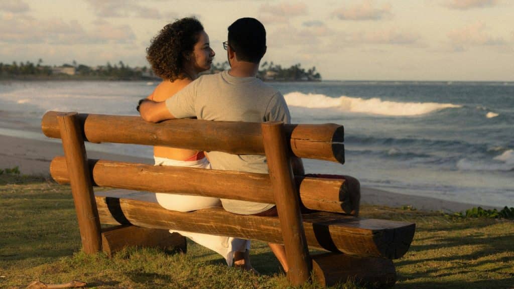 A couple sits on a wooden bench at the beach looking out at the ocean waves.