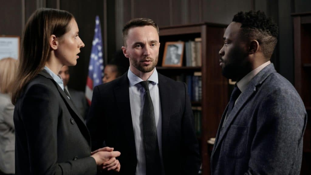 Three people in professional attire stand together in a room with a flag and bookshelves.