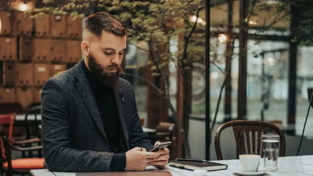 A man with a beard and suit jacket uses a smartphone at a white outdoor table.
