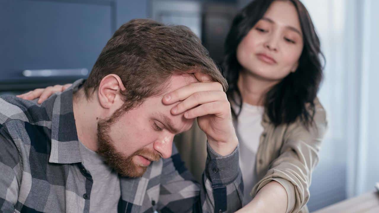A stressed man holding his head while a woman comforts him in the background.