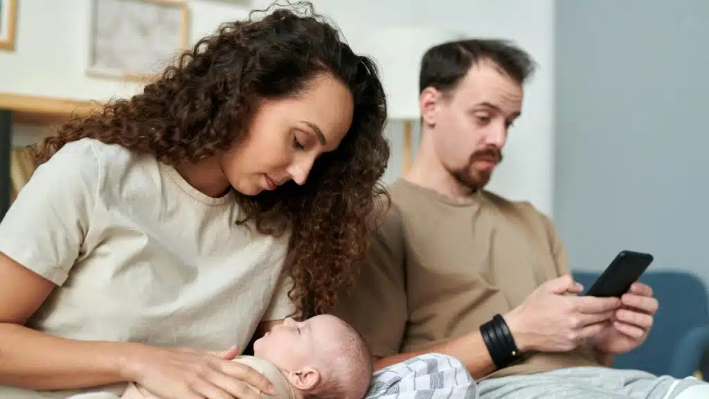 A woman looks at a baby while a man sits nearby looking at his phone.
