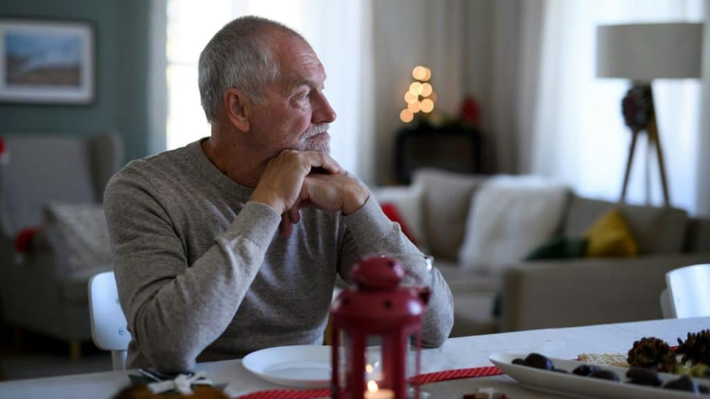 An older man sits at a table with his chin on his hands, looking sideways.