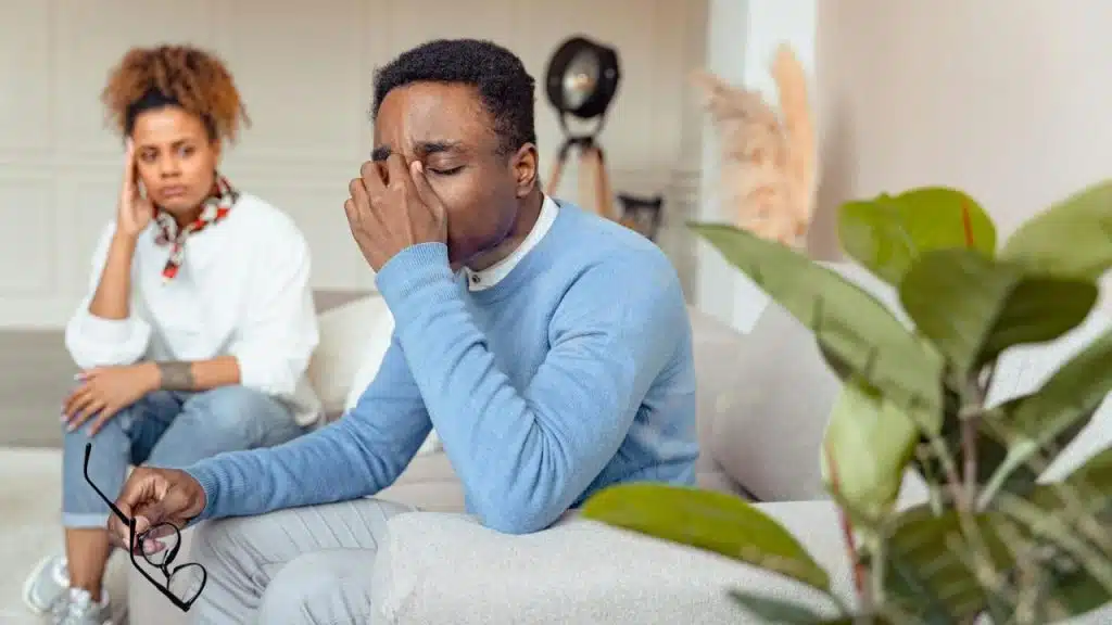 A man in a blue sweater pinches his nose while a woman sits behind him.