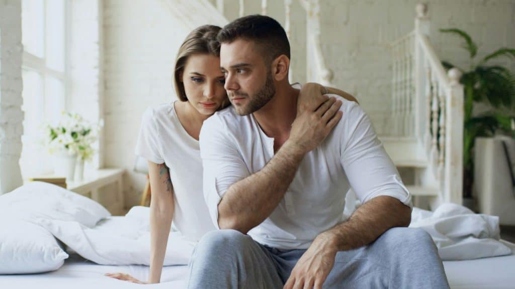 A man and woman in white shirts sit on a bed looking away with somber expressions.
