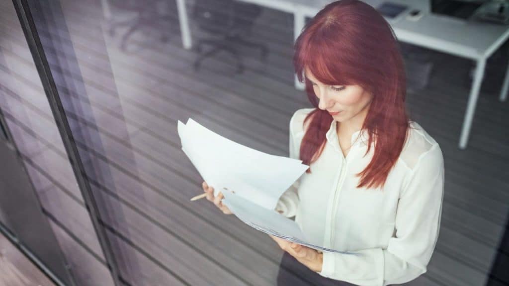 A woman with red hair wearing a white blouse looks down at papers in her hands.