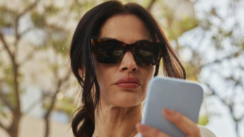 A woman wearing sunglasses looks at her smartphone outdoors.