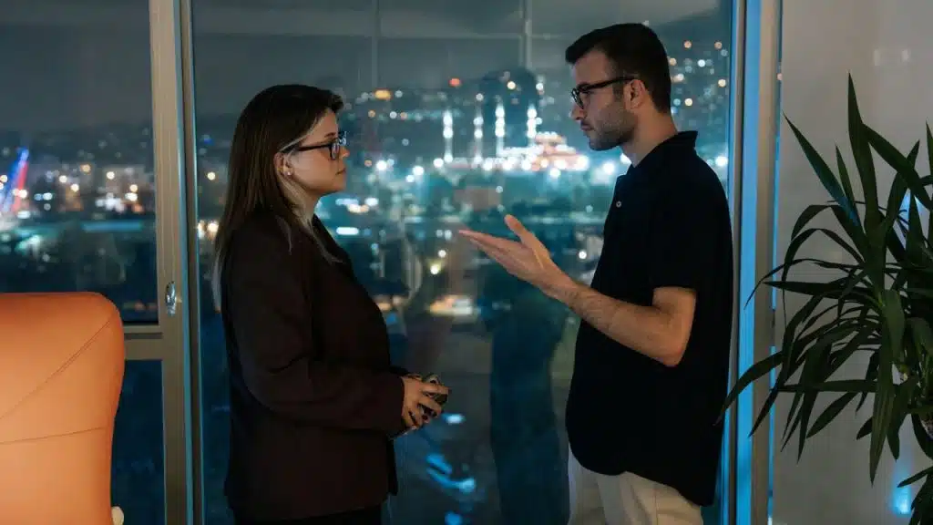 A man and woman wearing glasses stand by a window overlooking a glowing nighttime city.