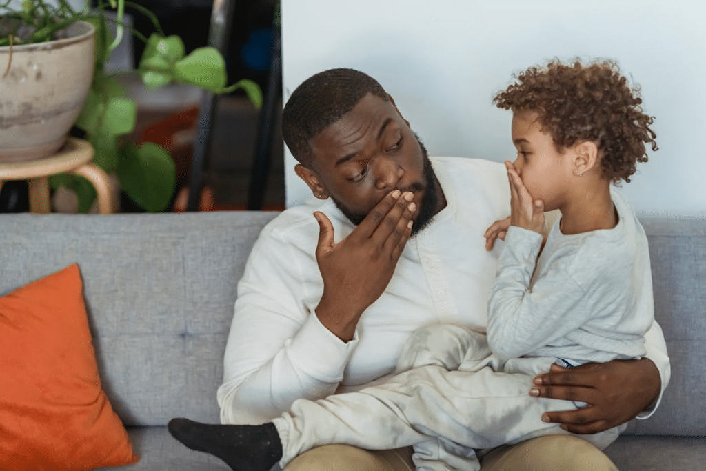 Black father with son covering mouth with hand on sofa