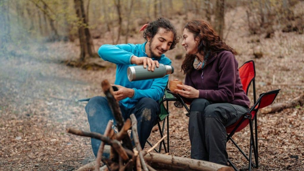 A man in a blue jacket pours a drink for a woman near a campfire.
