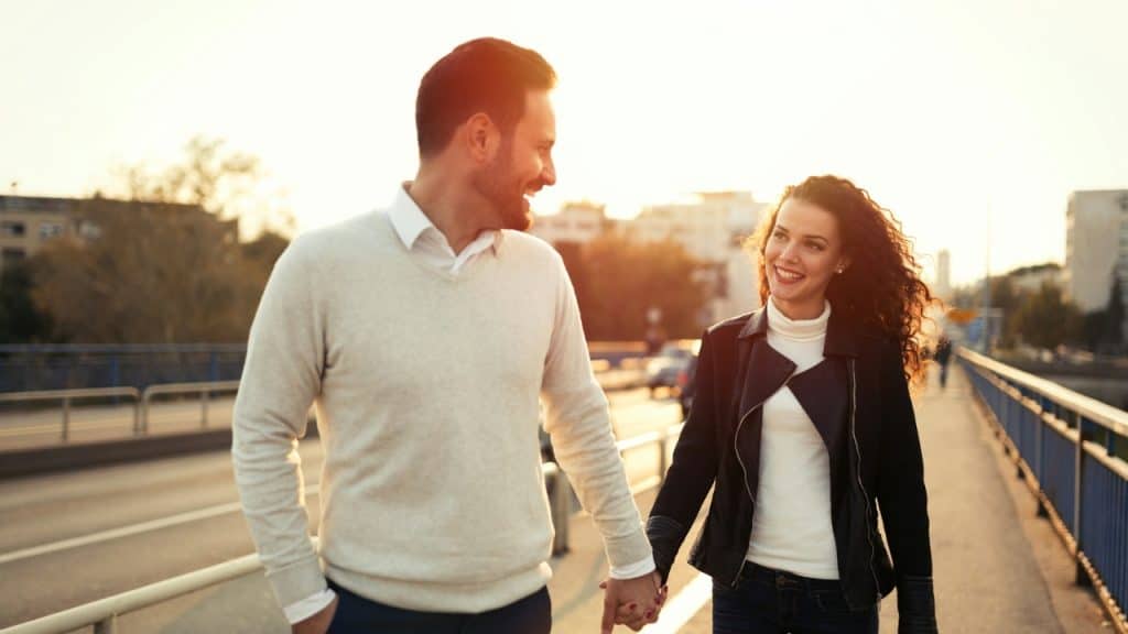 A smiling man and woman hold hands while walking outdoors on a sunlit bridge.