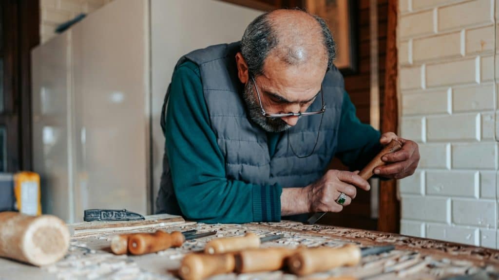 An older man with glasses leans over a wooden table while using a carving tool.
