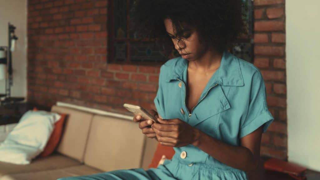 A woman sits on a couch and looks down at her phone in a brick-walled room.