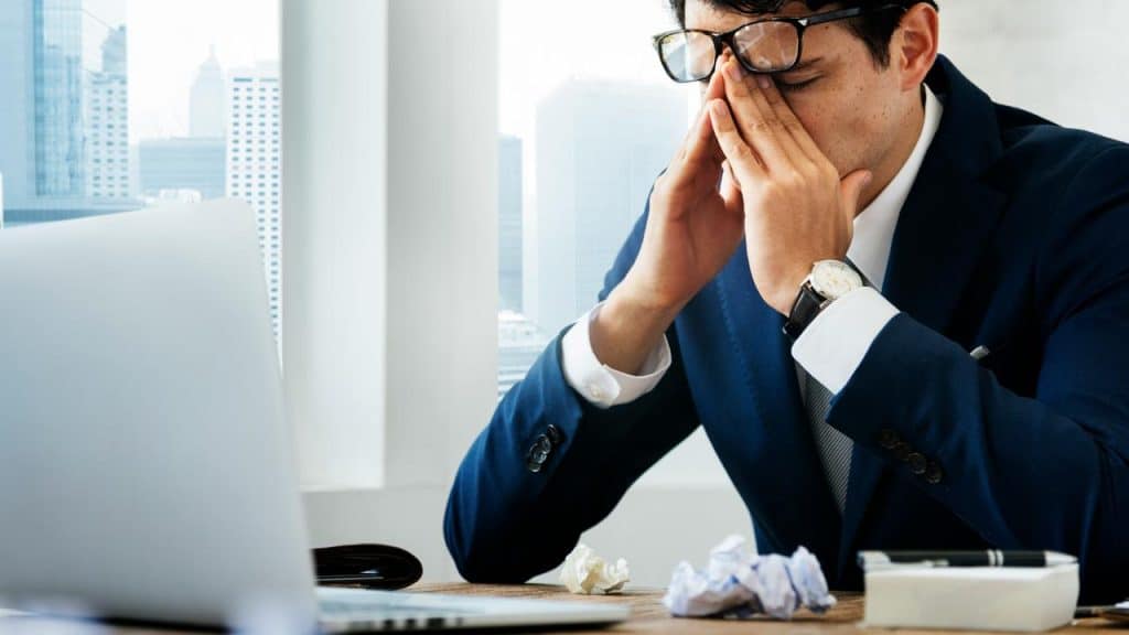 A stressed man in a suit rubbing his face at a desk with a laptop.