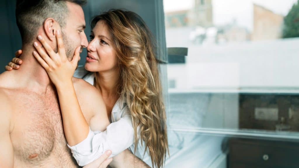 A woman with long blonde hair sits close to a shirtless man, holding his face tenderly as they gaze at one another in a bright room with a large window.
