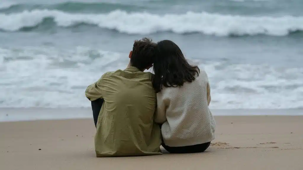 A rearview shot of a couple sitting close together on a sandy beach, their heads touching as they watch the white waves roll in under a grey, overcast sky.