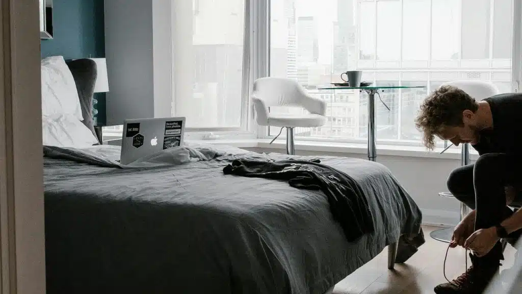 A man ties his shoes in a bedroom with a laptop on the bed.