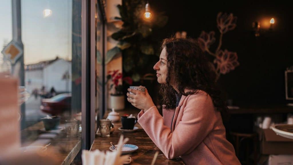 A woman in a pink coat sits by a café window holding a cup and gazing outside.