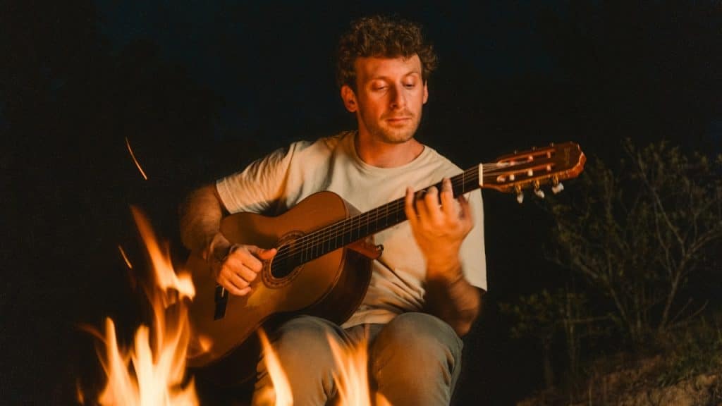 A man sits outdoors at night playing an acoustic guitar next to a bright campfire.
