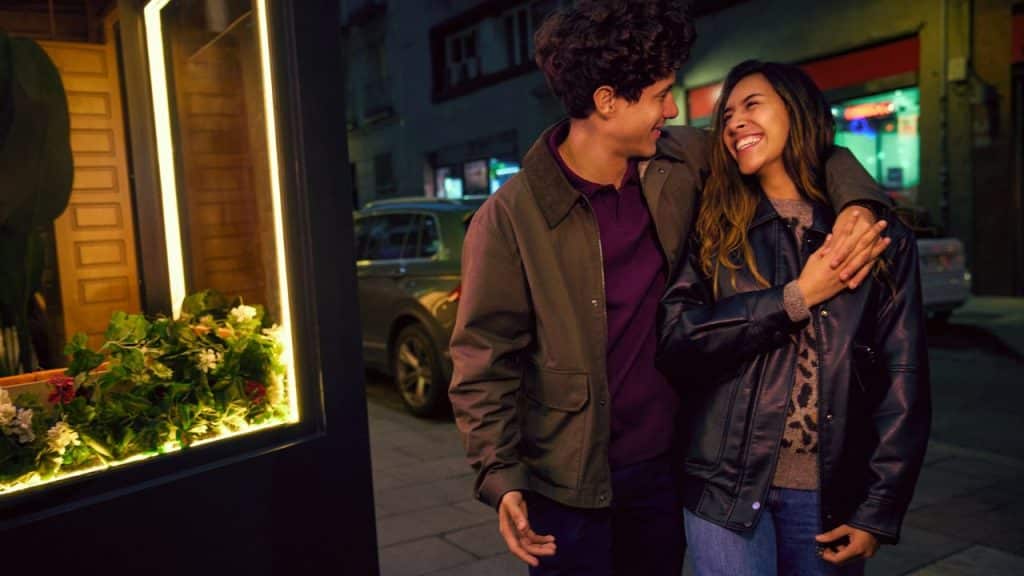 A man and woman walk at night, laughing with his arm around her shoulder.