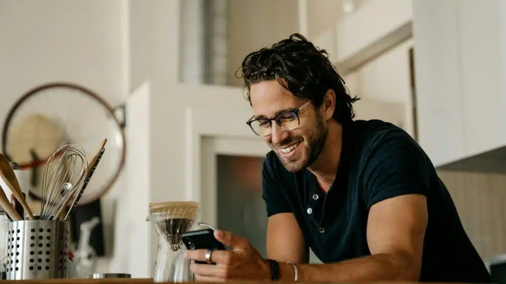 A man with glasses smiles while looking at a smartphone in a bright kitchen setting.