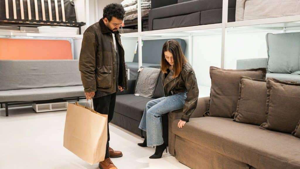 A man holding a large shopping bag watches a woman sit on a brown sofa.