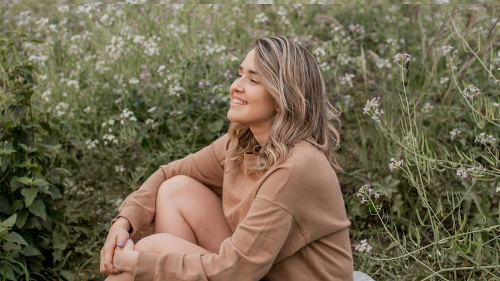 A woman smiling and sitting in a field while holding her legs with her arms.