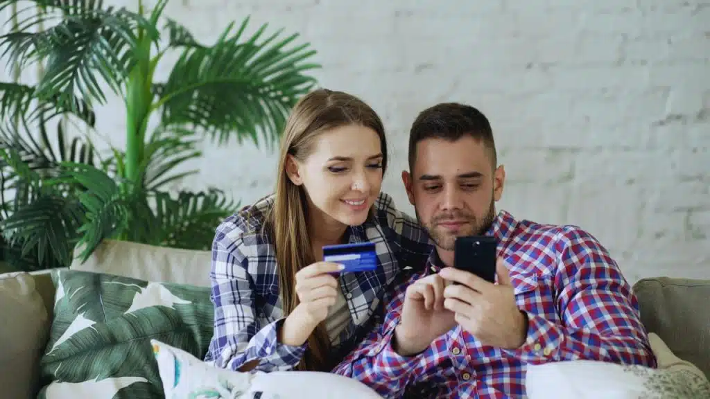 A smiling couple sitting on a couch, shopping online with a credit card and smartphone.