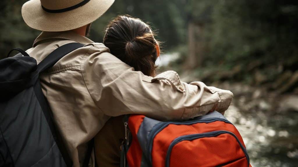 A couple with backpacks hugs while looking at a forest stream.