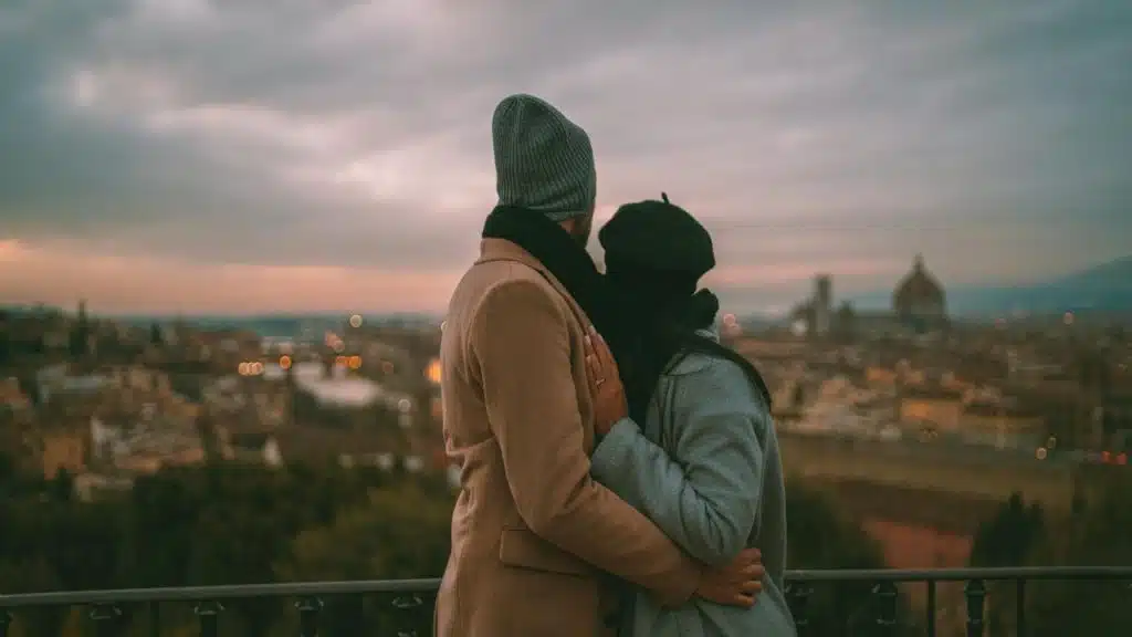 A couple embraces while looking out over a city at sunset.