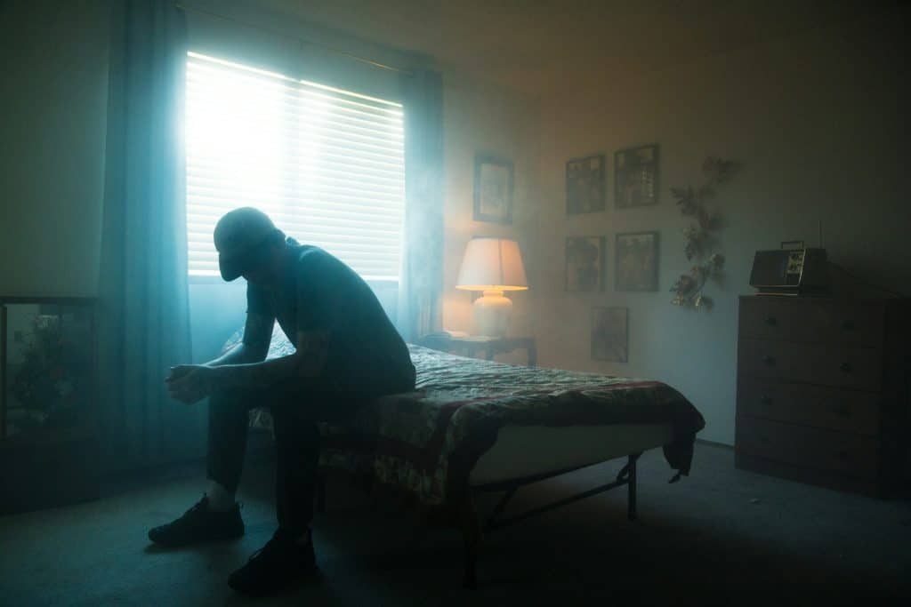 A man sitting on a bed in a dimly lit room and appears distraught.