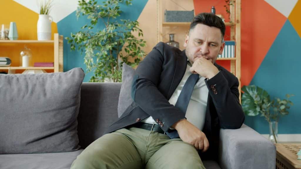A man in a suit and tie sits on a grey couch, resting his chin.